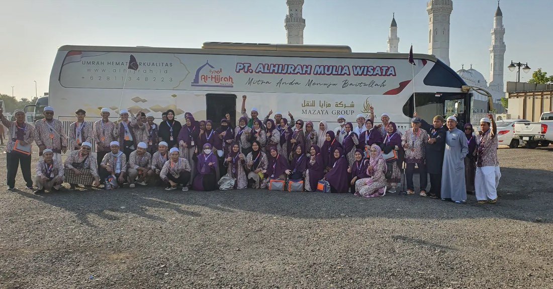 Rombongan besar jamaah berseragam ungu berfoto di depan bus dengan spanduk PT Al Hijrah Mulia Wisata